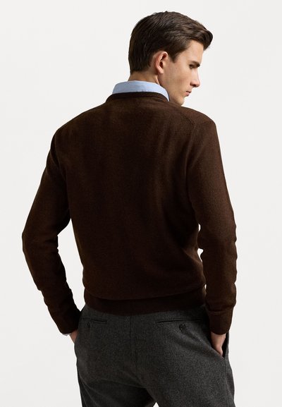 Suéter de punto marrón oscuro con cuello redondo, puños y bajos de canalé, llevado sobre una camisa de cuello azul claro. Los pantalones grises son parcialmente visibles.