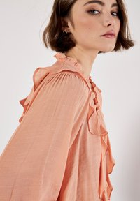 Blusa de color durazno hecha de una tela ligera, con acentos de volantes en el escote y las mangas, y un corte holgado.