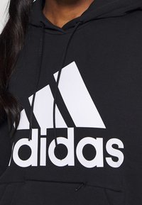 Černá mikina s kapucí vyrobená z měkké látky, s velkým bílým logem Adidas na hrudi a šňůrkami na kapuci.