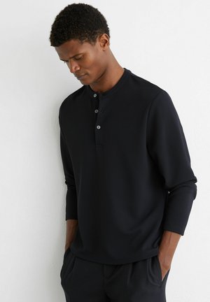 Mężczyzna w czarnej koszulce henley z długim rękawem i trzema guzikami, stojący z rękami w kieszeniach, patrzący w dół na tle białej ściany.