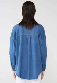 Camicia di jeans in blu chiaro, con vestibilità ampia, maniche lunghe, orlo curvo e bordo sfilacciato nella parte inferiore. Design minimalista.