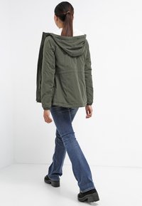 Oliwkowa, kapturowa kurtka z futerkiem z syntetycznego materiału, z zamkiem błyskawicznym i bocznymi kieszeniami, połączona z rozszerzanymi niebieskimi dżinsami i czarnymi butami na platformie.