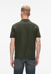Khaki zaļš polo krekls ar klasisku apkakli, īsām piedurknēm un gludu tekstūru. Valkāts kopā ar ziliem džinsiem, skats no aizmugures.