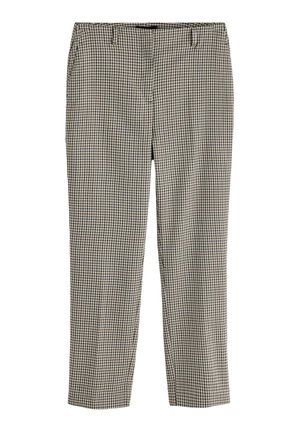 Frau trägt einen schwarz-weißen karierten Hosenanzug, schwarze Sonnenbrille, schwarze Sandalen, trägt eine schwarze Handtasche und geht drinnen an schlichten Wänden entlang.