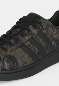 Χαμηλό sneaker με μαύρα κορδόνια, μαύρη σόλα και επάνω μέρος με καμουφλάζ μοτίβο που έχει τρεις μαύρες ρίγες στο πλάι.