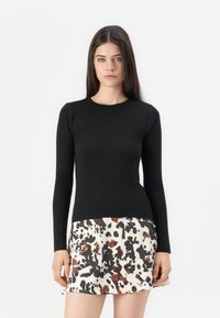 Donna con lunghi capelli castani indossa un top aderente nero a maniche lunghe e una gonna bianca con macchie astratte nere e marroni.
