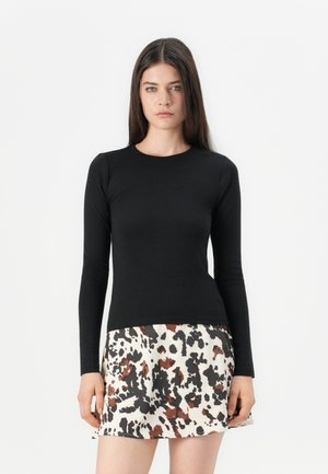 Woman with long brown hair wears a fitted black long-sleeve top and a white skirt with black and brown abstract spots.