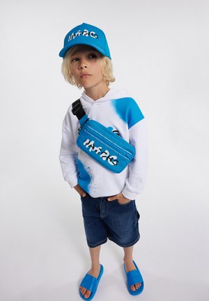 Felpa bianca con design spray blu, berretto blu, marsupio blu, pantaloni shorts in denim e sandali blu. Outfit casual con accenti di colori brillanti.