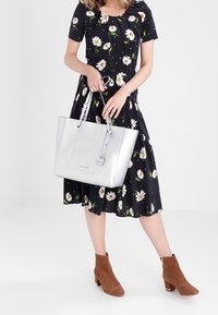 Srebrna torba na ramię o teksturowanej powierzchni, z podwójnymi uchwytami i zawieszką. Noszona z czarną sukienką w kwiaty oraz brązowymi botkami na obcasie.