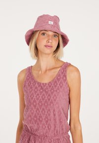 Chapeau seau en tissu rose poussiéreux texturé avec un patch logo blanc. Porté avec un haut sans manches assorti présentant un motif géométrique.