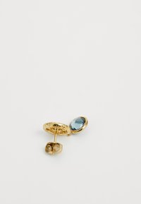 Boucles d'oreilles en métal doré avec une base circulaire texturée et une pierre précieuse ovale bleu clair. Le fermoir à tige est relié à un bouchon en caoutchouc transparent.