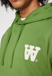 Green hoodie with a textured white embroidered logo and drawstrings. Material appears soft with a smooth finish.