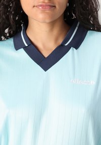 Lyseblå polo skjorte med vertikale striper, med navy krage og hvite detaljer samt en brodert "ellesse"-logo på brystet.