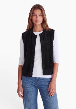 Frau trägt eine schwarze, flauschige Weste mit Knöpfen über einem weißen langärmeligen Hemd und blauen Jeans und steht vor einem schlichten weißen Hintergrund.