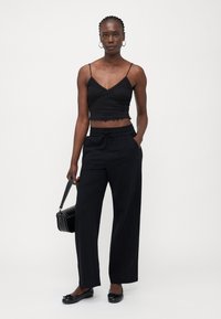 Svart cropped topp med blonderstruktur og bølget kant, sammen med løse svarte bukser. Modellen holder en svart håndveske og har på seg flate sko.