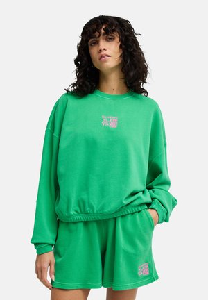 Junge Frau mit lockigem dunklem Haar, trägt einen hellgrünen Pullover und passende Shorts mit pinken Blumenlogos, Hände in den Taschen, neutraler Hintergrund.