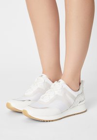 Scarpe sportive bianche caratterizzate da materiali in rete e pelle, chiusura con lacci, un accento argentato testurizzato e una suola in gomma di colore caramello.