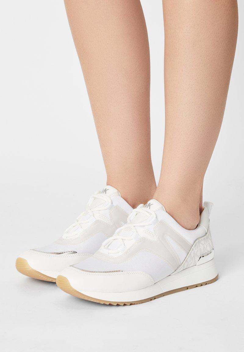 Scarpe sportive bianche caratterizzate da materiali in rete e pelle, chiusura con lacci, un accento argentato testurizzato e una suola in gomma di colore caramello.