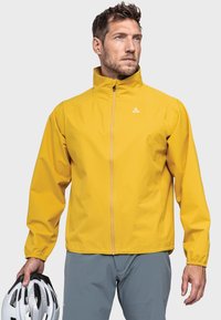 Gelbe Outdoor-Jacke mit hohem Kragen, Reißverschluss vorne und elastischen Bündchen. Glatte Textur, leichtes Material. Modell hält einen weißen Helm.