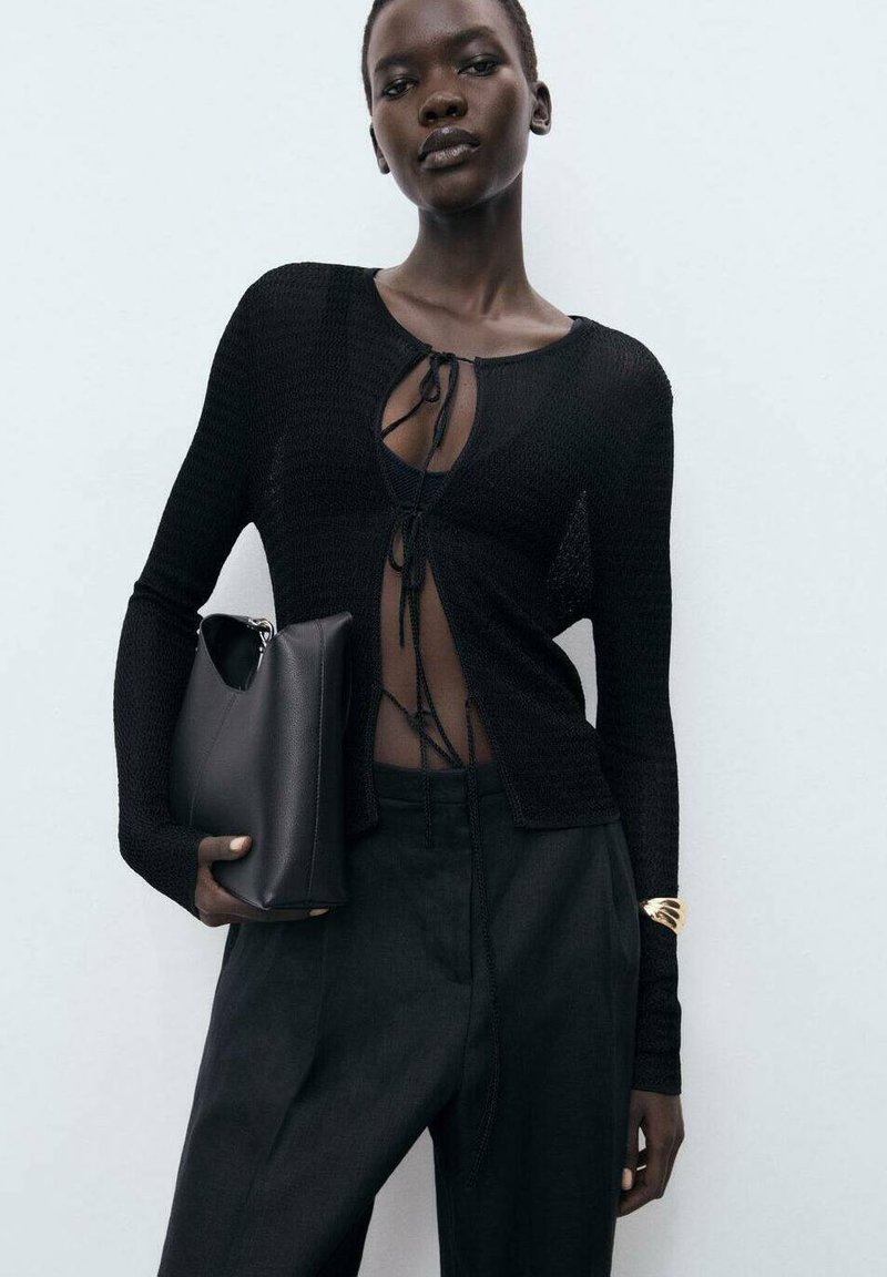 Zwarte gebreide top met lange mouwen en gevlochten voorkant, gecombineerd met op maat gemaakte zwarte broek en een gestructureerde zwarte clutch. Gladde textuur, minimalistisch ontwerp.