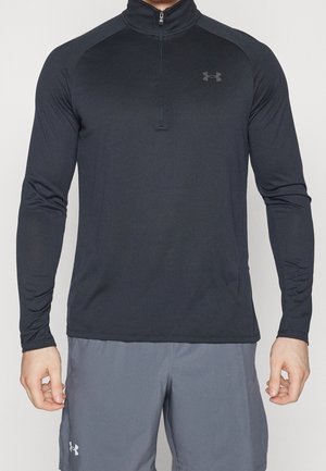 Mężczyzna ubrany w czarną, sportową koszulkę z długim rękawem i suwakiem oraz szare szorty, z logo Under Armour na piersi koszulki i na nodze szortów.