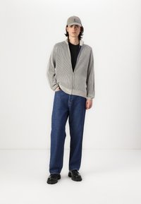 Grå tekstureret lynlås trøje, sort skjorte under, blå løstsiddende jeans og sorte sko. En beige kasket fuldender outfittet.