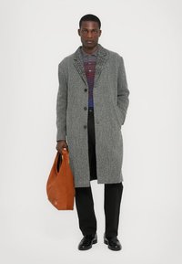 Szürke houndstooth mintás kabát, egygombos, két gombbal, csíkos póló felett viselve. A modell egy narancssárga táskával a kezében áll, fekete nadrágot és cipőt visel.