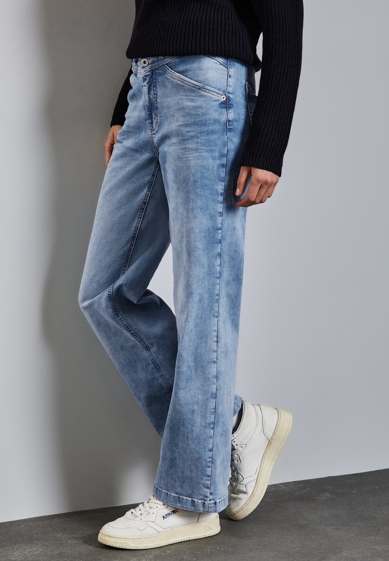 Vaqueros de pierna ancha en un tono azul claro de denim con una textura desgastada. Cuenta con una cremallera y botón en la parte delantera, pasadores para cinturón y bolsillos laterales. Combinados con zapatillas blancas.