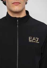 Svart zip-up jacka med hög krage, med en guld broderad EA7-logotyp på bröstet och en mjuk, lätt tygstruktur.