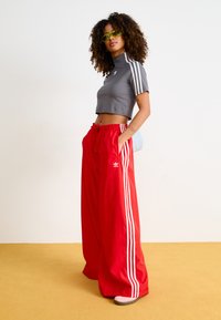 Haut de sport gris cropped avec col haut et trois rayures blanches. Pantalons larges rouges avec rayures blanches et logo. Le modèle porte des lunettes de soleil.