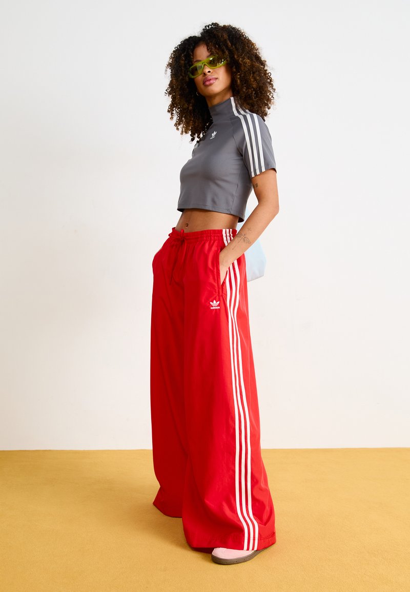 Haut de sport gris cropped avec col haut et trois rayures blanches. Pantalons larges rouges avec rayures blanches et logo. Le modèle porte des lunettes de soleil.