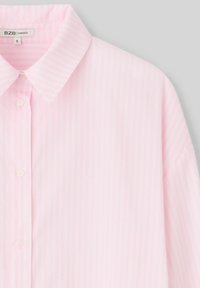 Blouse surdimensionnée rose clair avec des rayures verticales blanches, un col classique et une fermeture à boutons à l'avant avec des boutons blancs. Texture lisse.