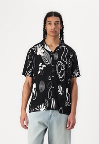 Kortermet svart skjorte med hvite abstrakte mønstre og former. Knappelukking foran; ledig passform; lett stoff. Båret med lyseblå jeans.