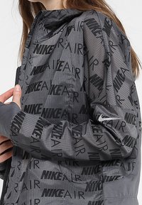 Coupe-vent Nike, présentant un tissu gris avec un motif en relief "NIKE AIR" en noir. Comprend une fermeture éclair et des trous pour les pouces aux poignets.