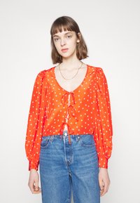 Blusa vermelha com bolinhas brancas, decote V profundo, frente com laço ajustável, mangas bufantes longas, combinada com jeans azuis de cintura alta.
