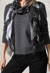 Femme portant un pull gris foncé avec poignets et ourlet côtelés, un pantalon noir et une écharpe noire à motif avec des accents bleu clair et violet.