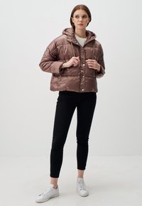Braune gesteppte Daunenjacke mit Kapuze, ausgestattet mit einem Reißverschluss und Druckknöpfen, über einem weißen Hemd präsentiert und kombiniert mit schwarzen Hosen und weißen Sneakers.