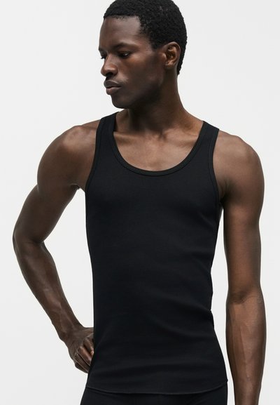 Αθλητικός άντρας με εφαρμοστή μαύρη ριμπ μπλούζα χωρίς μανίκια, κοιτάζοντας αριστερά με το ένα χέρι στη μέση, σε απλό φόντο.