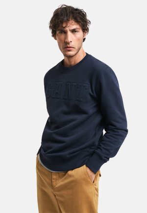 Sudadera azul marino hecha de una tela suave, con un logo "GANT" texturizado y elevado en la parte frontal, cuello redondo y puños de canalé.