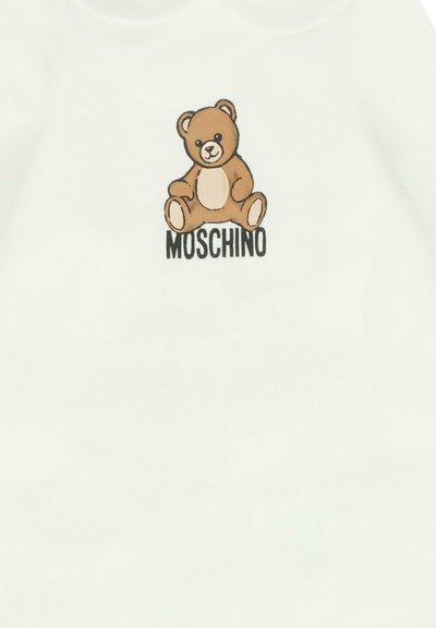 Un orsetto marrone seduto sopra il testo "MOSCHINO" su uno sfondo di tessuto bianco.