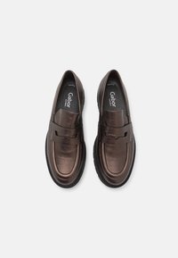 Mocassins en cuir marron avec une texture lisse, présentant une sangle proéminente sur le dessus et des semelles intérieures rembourrées pour le confort. Semelles en caoutchouc noir.