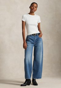 Kvinde iført hvid t-shirt med korte ærmer og rund hals, højtaljede blå jeans med brede ben og sort bælte samt sorte ankelstøvler, står med hånden i lommen.