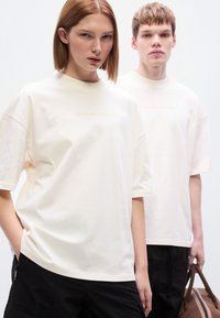 Βαμβακερές μπλούζες σε κρεμ χρώμα με κοντά μανίκια και χαλαρή εφαρμογή. Διαθέτουν ανάγλυφο κείμενο «BROKE SINCE MILANO» στο μπροστινό μέρος. Συνδυάζονται με σκούρο παντελόνι.