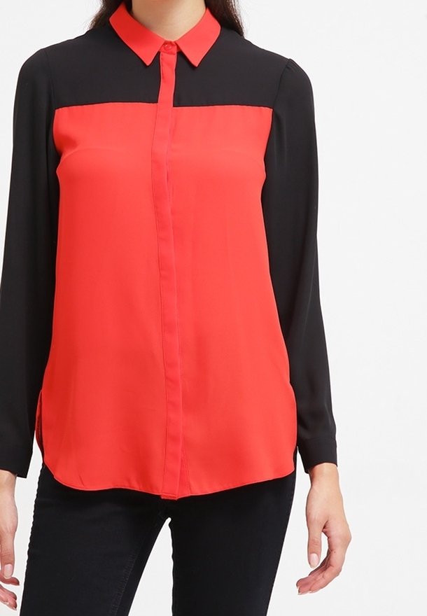 Femme portant un chemisier à manches longues avec un devant rouge et des manches et épaules noires, associé à un pantalon noir.