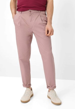 Mann trägt staubrosa Chinos mit hochgekrempelten Bündchen, ein burgunderfarbenes Hemd, das in die Hose gesteckt ist, und cremeweiße Sneaker mit Gummisohlen, steht mit den Händen in den Taschen.