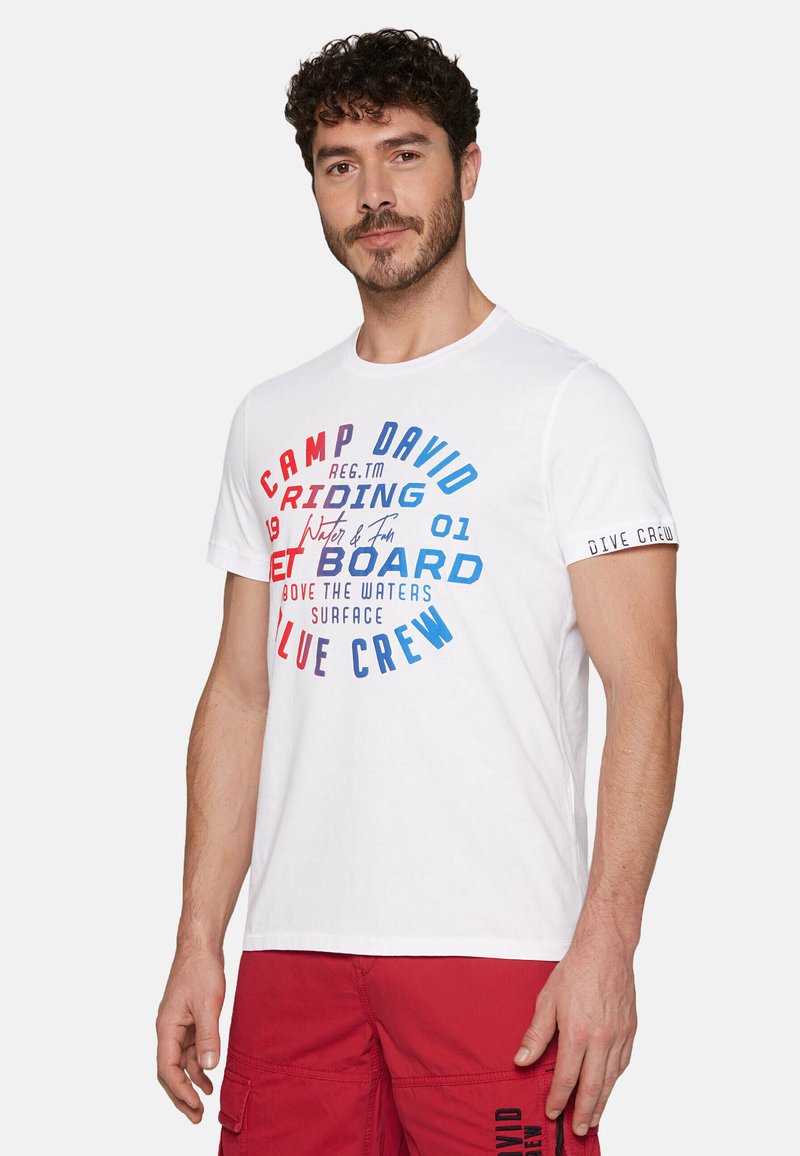 Bílý tričko s vícebarevnými textovými grafikami. Text zahrnuje "CAMP DAVID," "RIDE" a "BLUE CREW." Krátké rukávy s jemným akcentem.