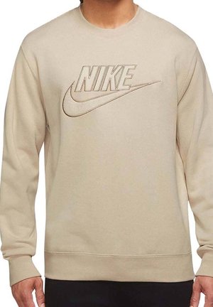 Beżowa bluza Nike z długim rękawem, z haftowanym logo na piersi, noszona przez osobę z rozluźnionymi rękami u boków.
