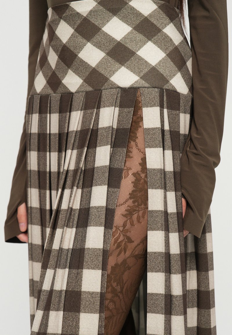 Falda a cuadros marrón y crema con pliegues y una abertura alta en el muslo, que revela una capa con patrón de encaje floral por debajo. Top de mangas largas en marrón oscuro.