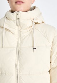 Puffer jacket in light beige, featuring a zip-up front, adjustable hood with drawstrings, and subtle logo detail on the chest.