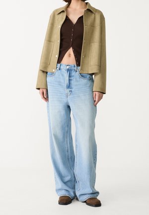 Femme portant un jean bleu clair à jambes larges, un top court marron boutonné, une veste vert olive et des chaussures marron, debout sur un fond uni.
